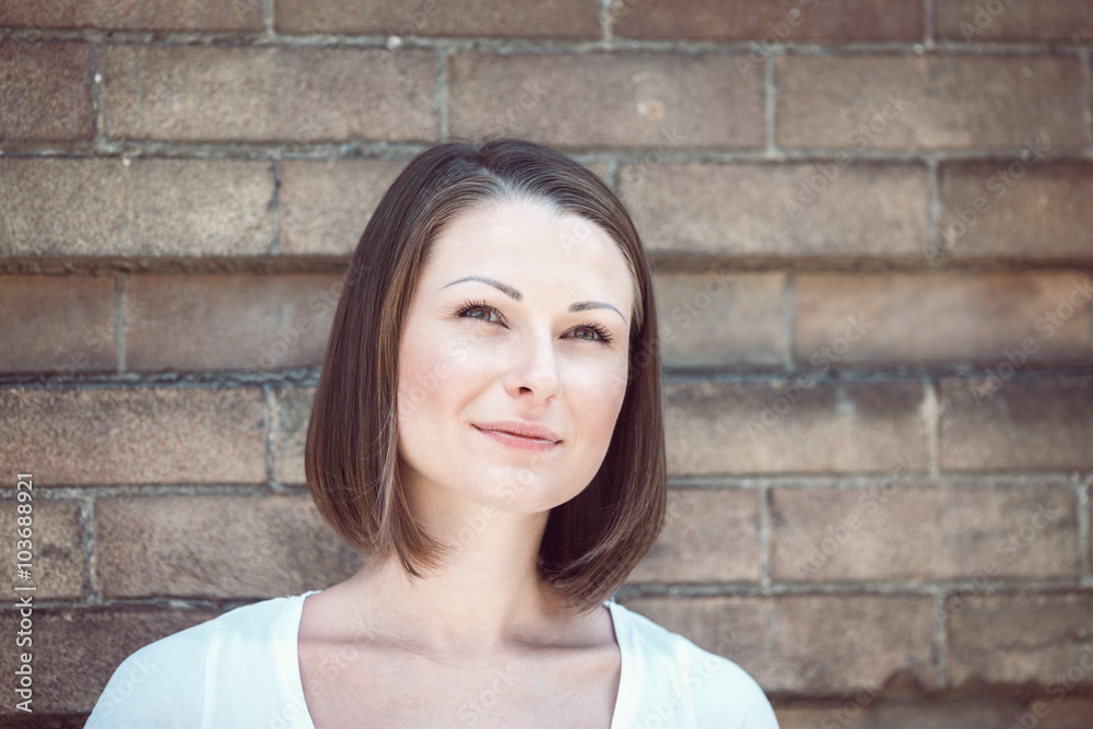 Close-up portrait of beautiful innocent Caucasian adult girl woman with long hair, bob style, hazel eyes, in white tshirt standing behind brick wall outside looking away