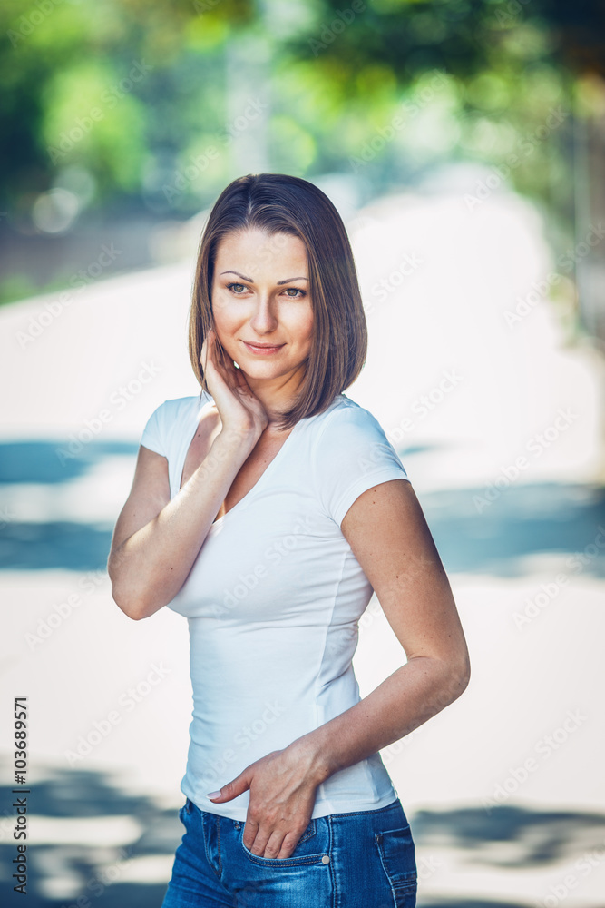 Portrait of beautiful innocent Caucasian adult girl woman with long hair,  bob style, hazel eyes, in white tshirt and blue jeans, standing in park  street outside looking away from camera Stock Photo