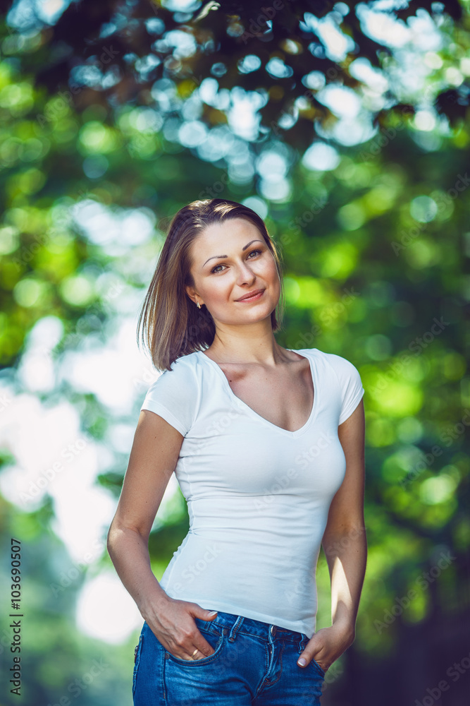 Portrait of beautiful innocent Caucasian adult girl woman with long hair, bob style, hazel eyes, in white tshirt and blue jeans, standing in park street outside looking in camera