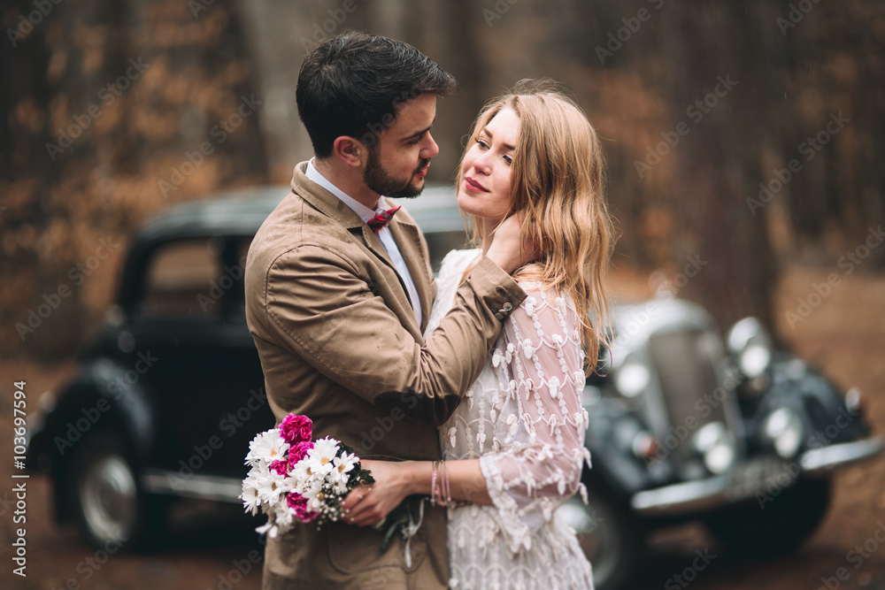 Obraz premium Gorgeous newlywed bride and groom posing in pine forest near retro car in their wedding day