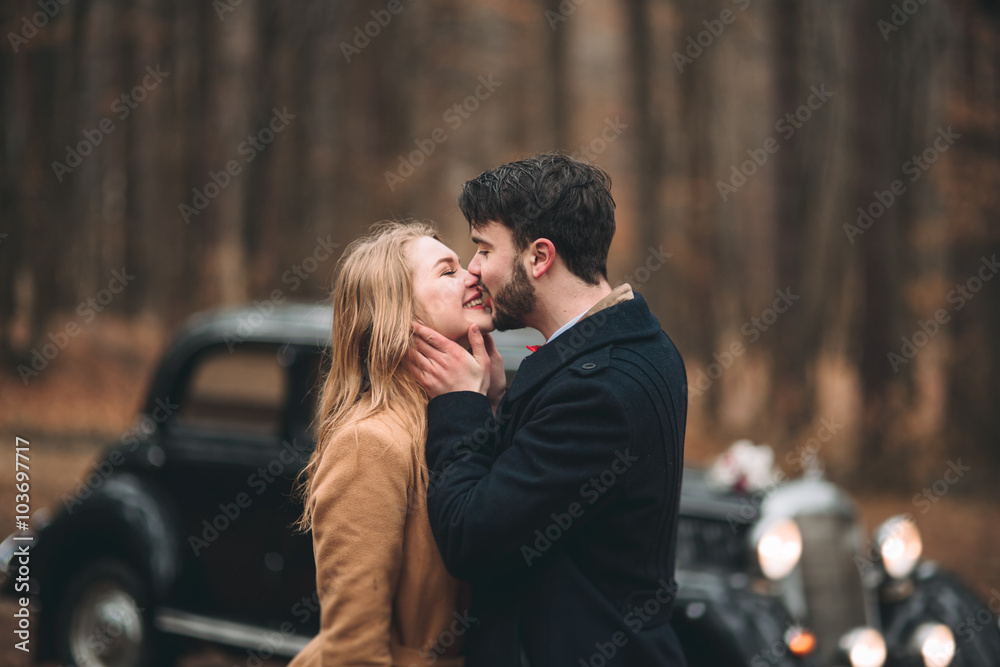 Gorgeous newlywed bride and groom posing in pine forest near retro car in their wedding day Gorgeous newlywed bride and groom posing in pine forest near retro car in their wedding day