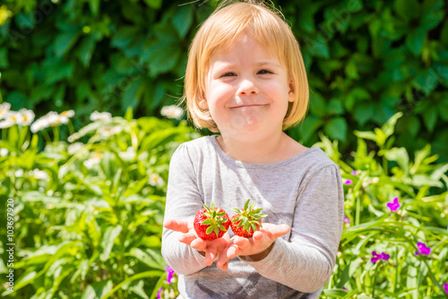 Smiling little girl holds in hands two strawberries. Sunny summer day on countryside village.