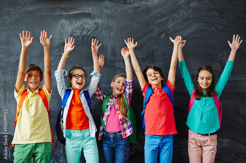 Ecstatic kids raising hands while standing by blackboard