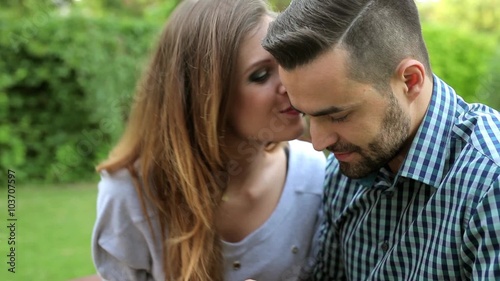 Couple sitting on the bench in the garden. Man is using phone and woman is kissing him