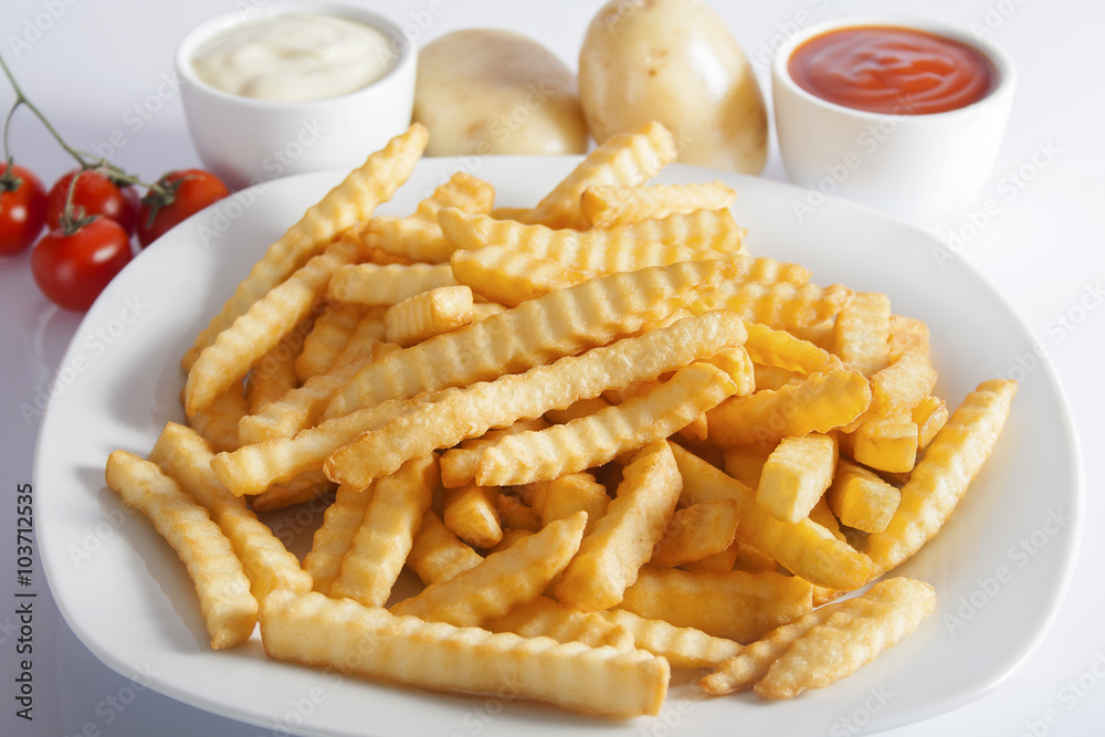 Portion of French fries (Crinkle-cut) deep fried, served on a white plate next to white bowls with mayonnaise and ketchup and fresh potato.