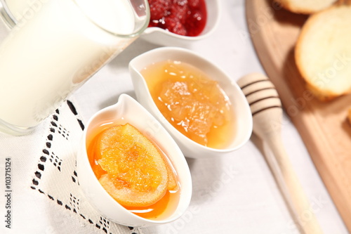 White bowls with homemade jams next to a glass of milk and sweet baked bun are on linen tablecloth on a rustic table