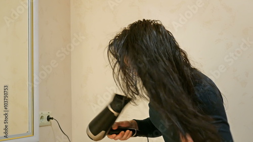 Beautiful brunette woman drying long black silky hair with hairdryer and smartens up in front of mirror in make-up dressing room. 4K UHD video footage. photo