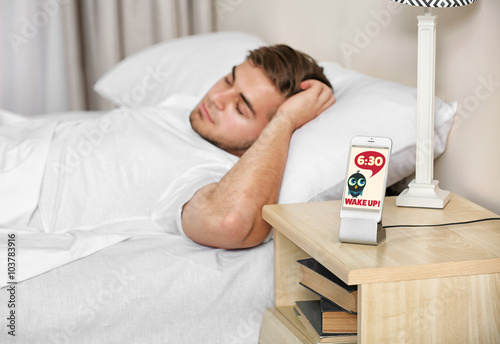 Beautiful young man waking up with mobile alarm clock photo