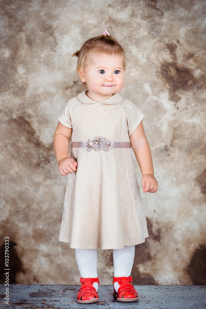 Adorable little girl with blond hair and plump cheeks wearing stylish beige dress and red shoes Adorable little girl with blond hair and plump cheeks wearing stylish beige dress and red shoes