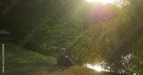 Man Sits On The Ground At The Lakeshore He Holds Laptop On His Knees And Looks At The Water Then Man Puts Backpack Under His Head And Leans Back