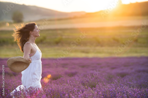 Fototapeta Naklejka Na Ścianę i Meble -  Beautiful young  healthy woman with a white dress running joyfully through a lavender field holding a straw hat under the rays of the setting sun.