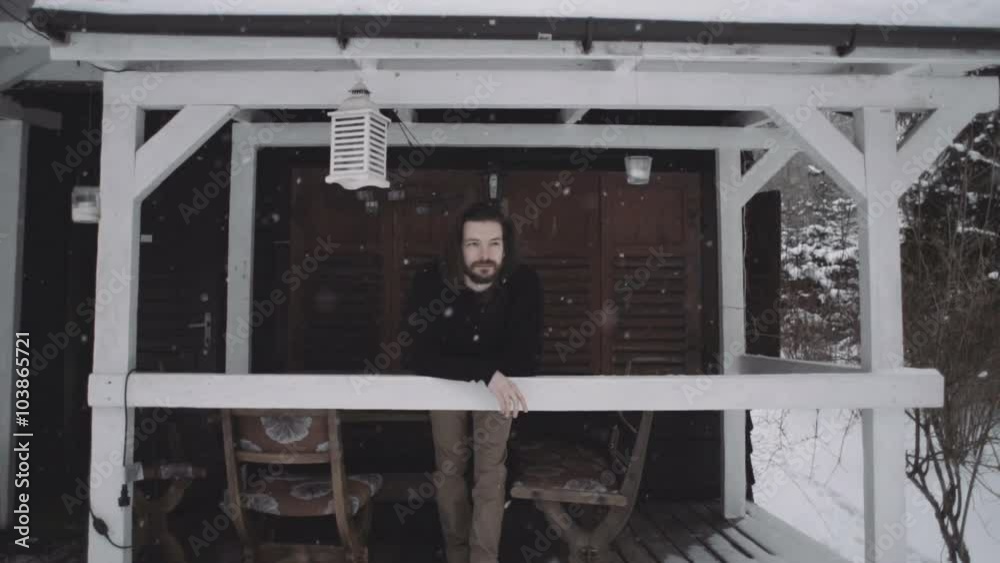 Handsome young man in black sweater standing on the wooden porch. Winter scene of pensive young man standing outdoors. Snowy background. Slow motion. Handsome young man in black sweater standing on the wooden porch. Winter scene of pensive young man standing outdoors. Snowy background. Slow motion.