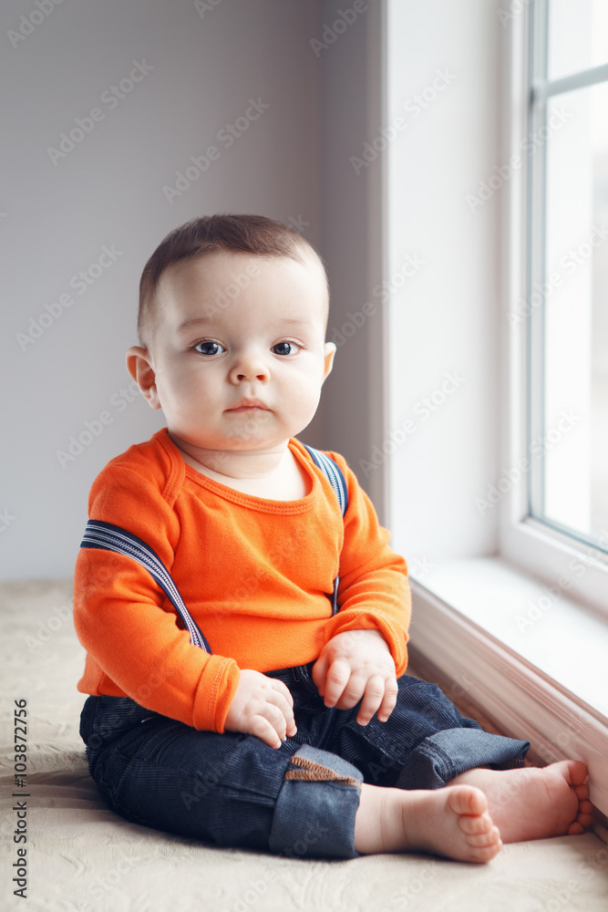 Portrait of cute adorable stylish Caucasian baby boy with black eyes in orange red shirt onesie, jeans with suspenders barefoot sitting on windowsill looking in camera, natural window light, lifestyle Portrait of cute adorable stylish Caucasian baby boy with black eyes in orange red shirt onesie, jeans with suspenders barefoot sitting on windowsill looking in camera, natural window light, lifestyle