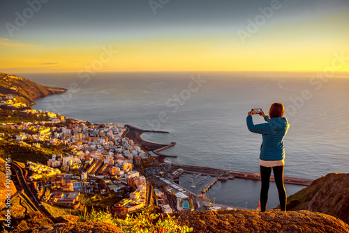 Young female traveler in blue jacket photographing Santa Cruz city standing on the top of the mountain on La Palma island in the morning photo