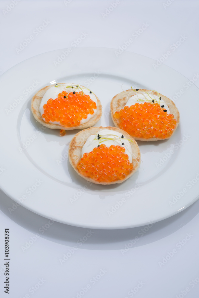 red fish caviar on pancakes with cream and leek sprouts. food on a white plate. space for text.