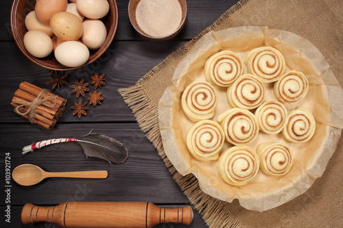 Raw cinnamon rolls prepared for roasting next to eggs and kitchen tools on a background of rustic table of black boards