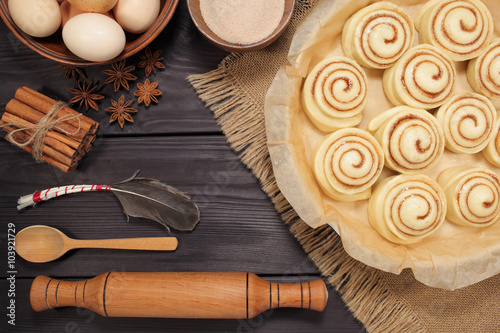 Raw cinnamon rolls prepared for roasting next to eggs and kitchen tools on a background of rustic table of black boards