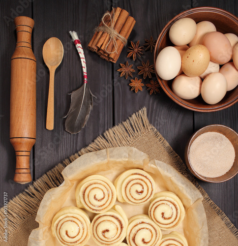 Raw cinnamon rolls prepared for roasting next to eggs and kitchen tools on a background of rustic table of black boards