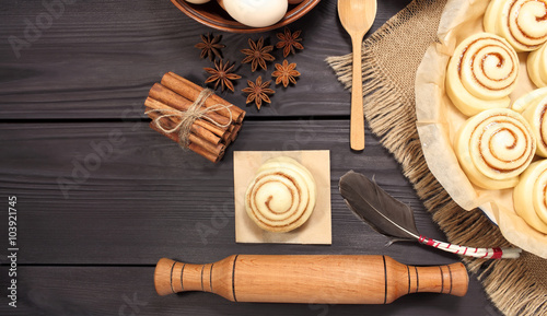 Raw cinnamon rolls prepared for roasting next to eggs and kitchen tools on a background of rustic table of black boards