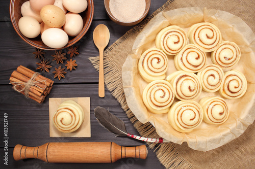 Raw cinnamon rolls prepared for roasting next to eggs and kitchen tools on a background of rustic table of black boards