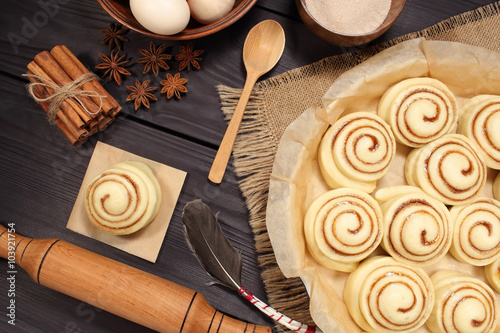 Raw cinnamon rolls prepared for roasting next to eggs and kitchen tools on a background of rustic table of black boards