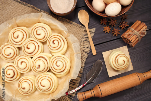Raw cinnamon rolls prepared for roasting next to eggs and kitchen tools on a background of rustic table of black boards