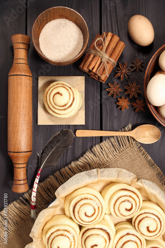 Raw cinnamon rolls prepared for roasting next to eggs and kitchen tools on a background of rustic table of black boards
