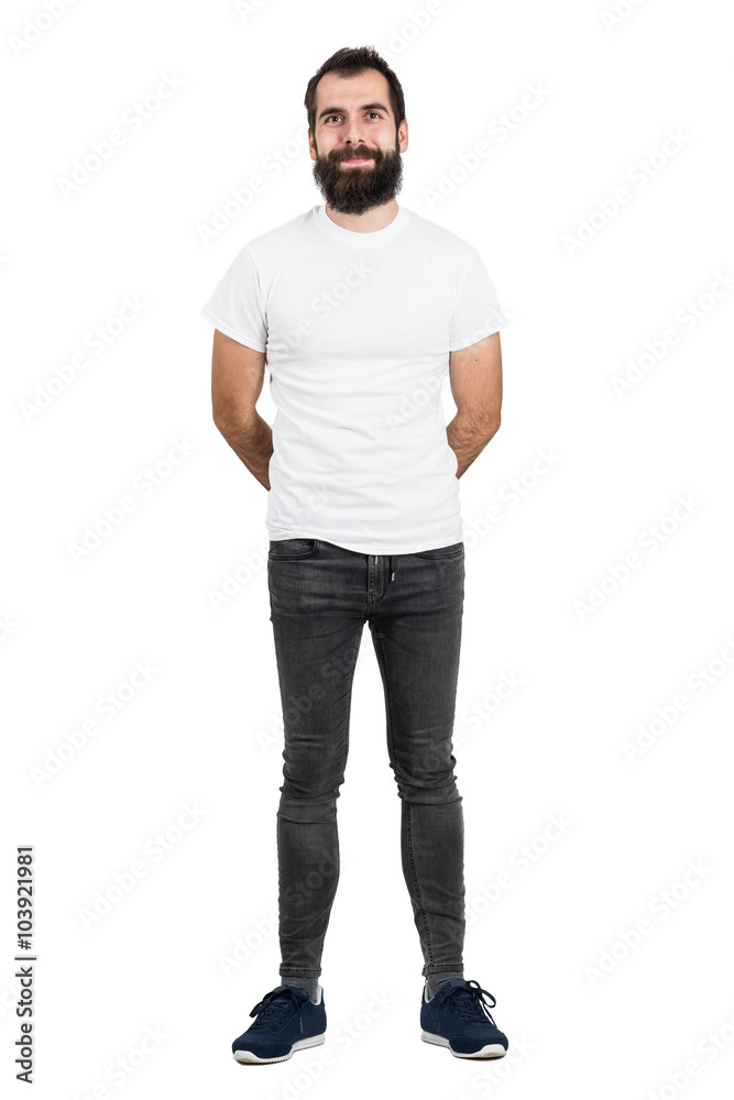 Happy bearded man with hands behind back in white t-shirt and tight jeans looking at camera. Full body length portrait isolated over white studio background. Happy bearded man with hands behind back in white t-shirt and tight jeans looking at camera. Full body length portrait isolated over white studio background.