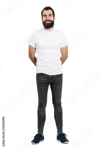 Happy bearded man with hands behind back in white t-shirt and tight jeans looking at camera. Full body length portrait isolated over white studio background. 