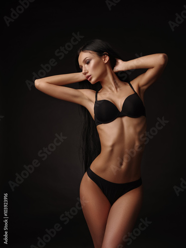 Slim beautiful model with dark straight hair and tanned skin in the black basic underwear collection is posing in the studio on the black background © maksimvostrikov