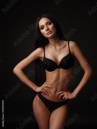 Young beautiful woman with dark straight hair, almond-shaped eyes and tanned skin in the black seamless underwear collection is posing in the studio on the black background © maksimvostrikov