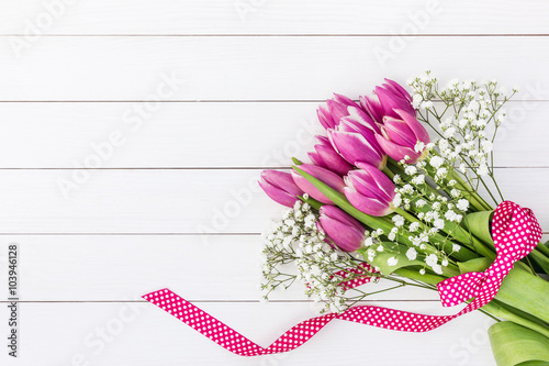 Fototapeta Naklejka Na Ścianę i Meble -  Bouquet of bright pink tulips and gipsophila decorated with ribbon on white wooden background. Top view, copy space