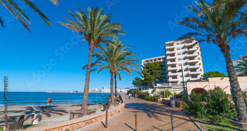 Mid morning sunshine in Ibiza. People walk along the beach near the city. Warm sunny day along water's edge. Ibiza, St Antoni de Portmany Balearic Islands, Spain. Row of palms lines the walkway.