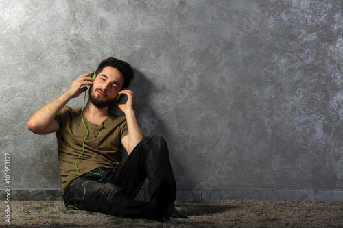 Young bearded man listens music with headphones sitting on grey wall background