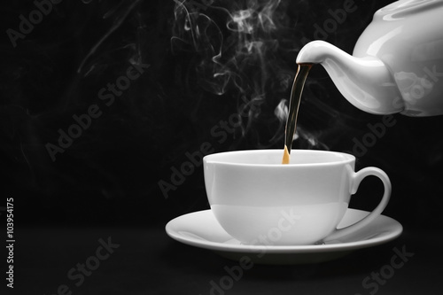 Pouring hot tea from a kettle into a cup on black background, close up