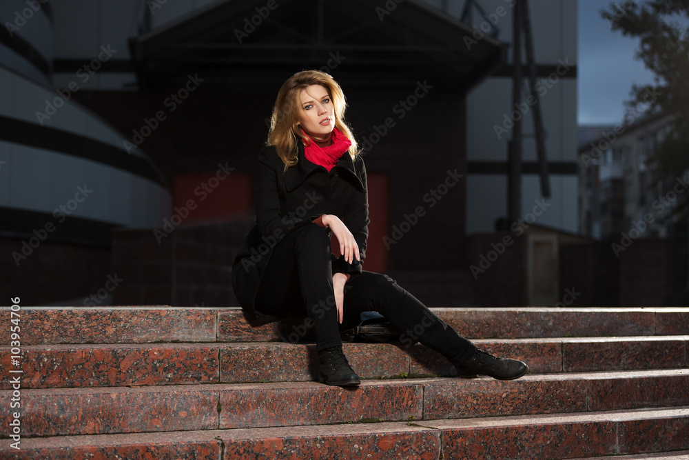 Fashion blond woman in black coat sitting on the steps