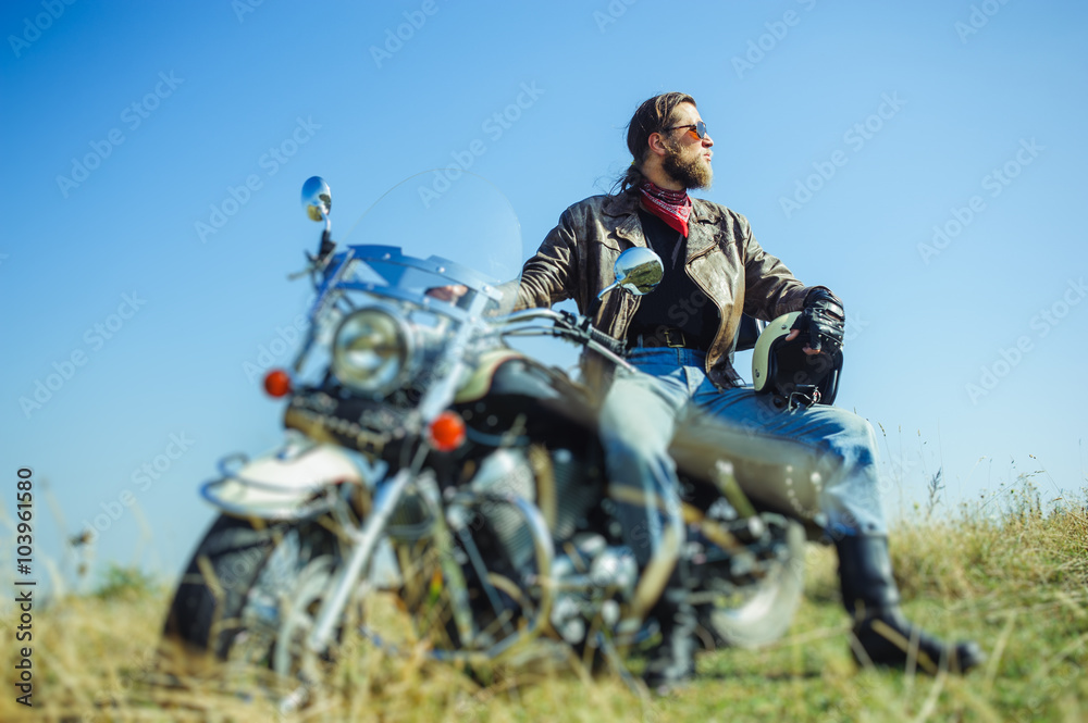 Portrait of a young man with beard sitting on his cruiser motorcycle and looking to the sun. Man is wearing leather jacket and blue jeans. Low point of view. Tilt shift lens blur effect Portrait of a young man with beard sitting on his cruiser motorcycle and looking to the sun. Man is wearing leather jacket and blue jeans. Low point of view. Tilt shift lens blur effect