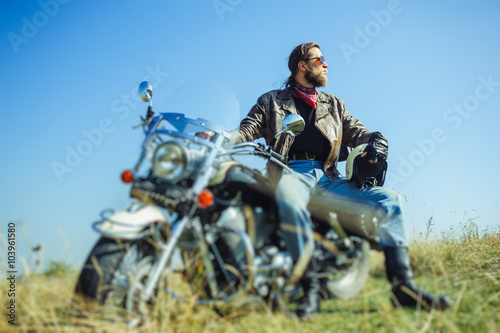 Portrait of a young man with beard sitting on his cruiser motorcycle and looking to the sun. Man is wearing leather jacket and blue jeans. Low point of view. Tilt shift lens blur effect