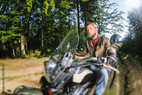 Biker with beard driving his cruiser motorcycle in the forest. Man is wearing leather jacket and blue jeans. Wide angle. Tilt shift lens blur effect