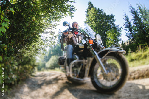 Young biker with beard driving his cruiser motorcycle in the forest. Man is wearing leather jacket and blue jeans. Low point of view. Tilt shift lens blur effect