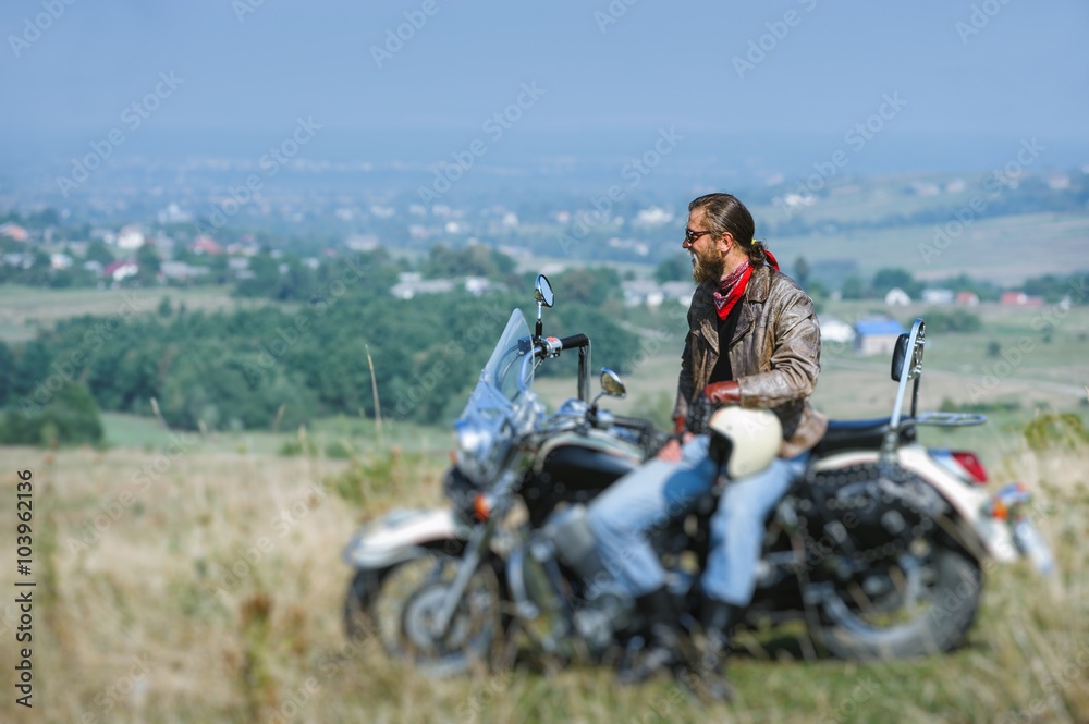 biker with beard wearing leather jacket and sunglasses sitting on his motorcycle on a sunny day, holding helmet. Horizontal picture. Tilt shift lens blur effect biker with beard wearing leather jacket and sunglasses sitting on his motorcycle on a sunny day, holding helmet. Horizontal picture. Tilt shift lens blur effect