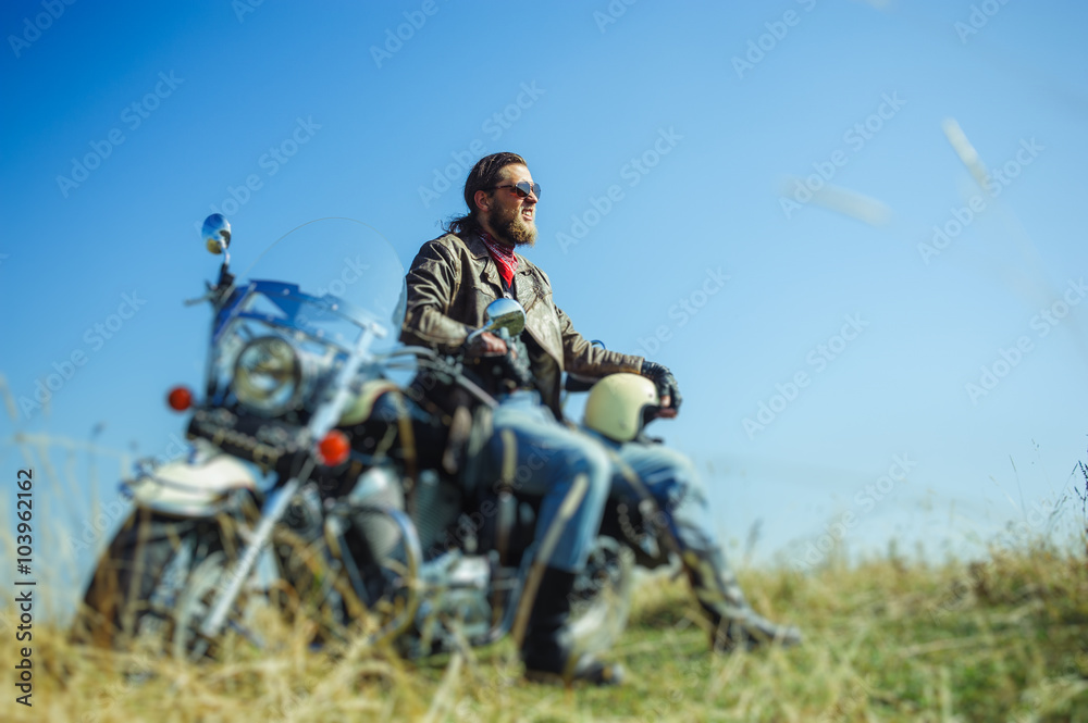 Portrait of a young man with beard sitting on his cruiser motorcycle and smilling. Man is wearing leather jacket and blue jeans. Low point of view. Tilt shift lens blur effect Portrait of a young man with beard sitting on his cruiser motorcycle and smilling. Man is wearing leather jacket and blue jeans. Low point of view. Tilt shift lens blur effect
