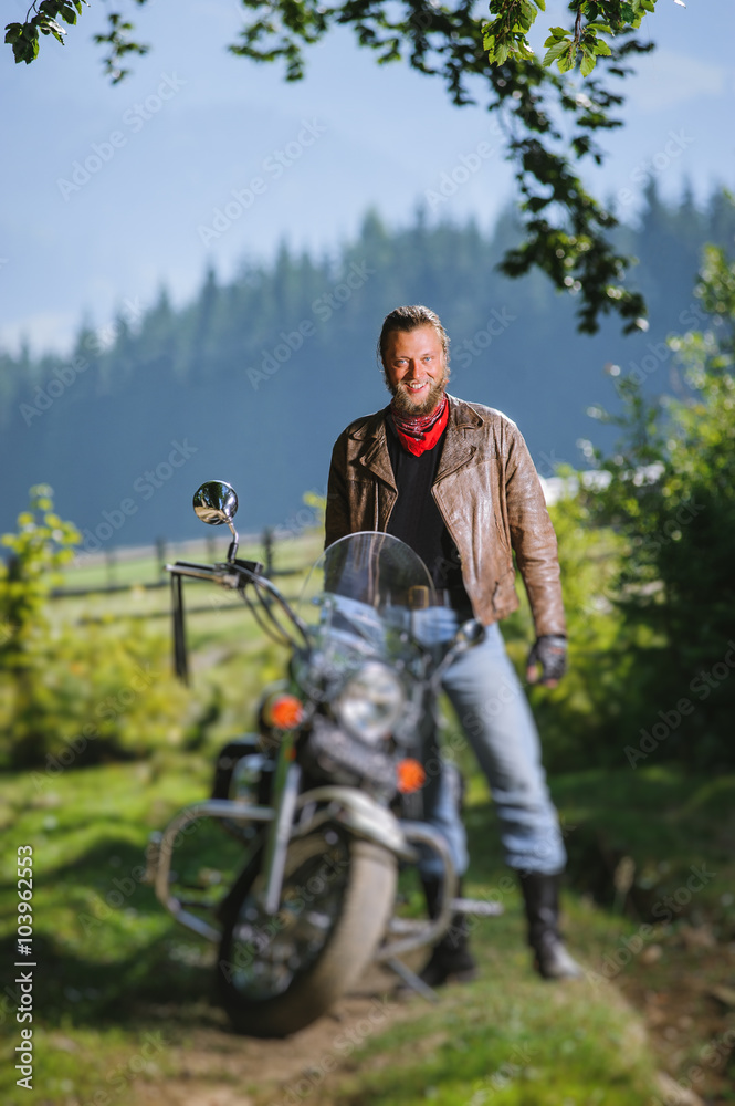 Naklejka premium Young handsome biker standing by his custom made cruiser motorcycle on a sunny day with forest on the background and smiling. vertical picture. Tilt shift lens blur effect