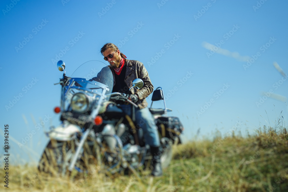 Portrait of a young biker with beard sitting on his cruiser motorcycle and looking to his bike. Man is wearing leather jacket and blue jeans. Low point of view. Tilt shift lens blur effect Portrait of a young biker with beard sitting on his cruiser motorcycle and looking to his bike. Man is wearing leather jacket and blue jeans. Low point of view. Tilt shift lens blur effect
