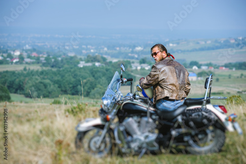 Brutal biker with beard wearing leather jacket and sitting on his motorcycle on a sunny day, holding helmet. Horizontal picture. View from the back. Looking to the camera. Tilt shift lens blur effect