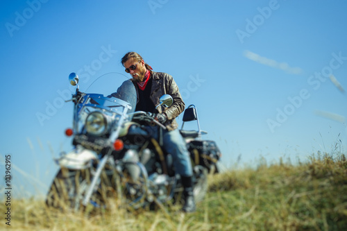 Portrait of a young biker with beard sitting on his cruiser motorcycle and looking to his bike. Man is wearing leather jacket and blue jeans. Low point of view. Tilt shift lens blur effect