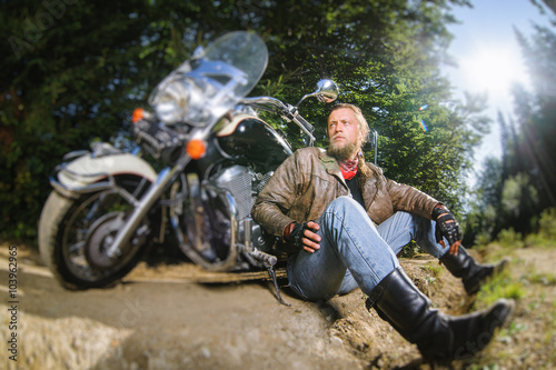 Bearded biker posing with his shiny stylish custom made cruiser motorbike. Biker is sitting on the ground near his bike wearing leather jacket, leather gloves and leather boots. tilt shift soft effect