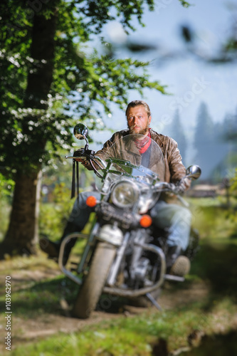 Handsome biker with beard driving his cruiser motorcycle in the forest and looking aheat. Man is wearing leather jacket and blue jeans. Tilt shift lens blur effect