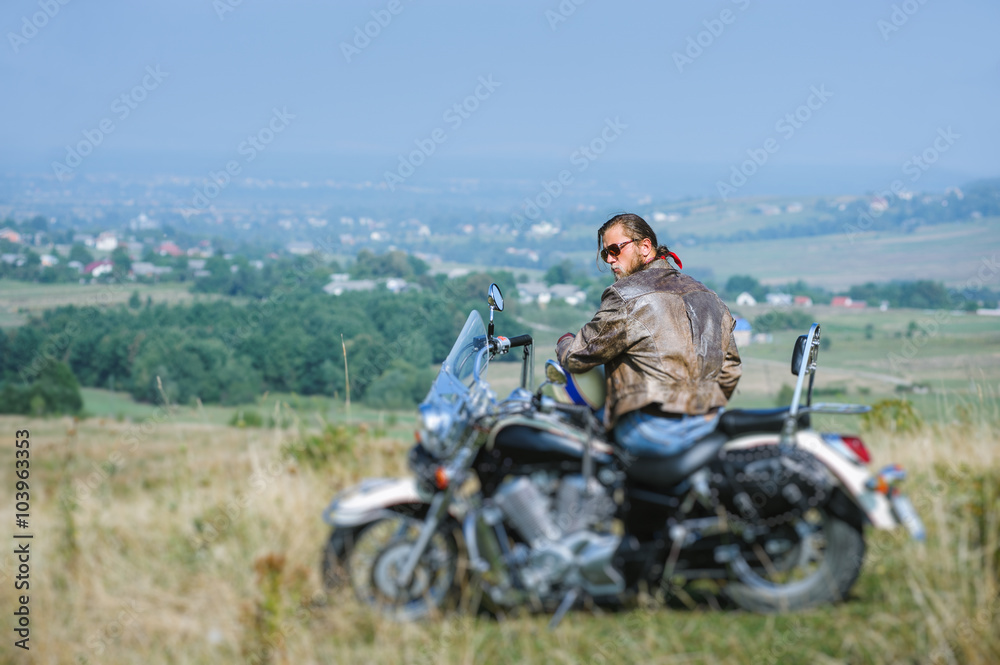 Naklejka premium Brutal biker with beard wearing leather jacket and sunglasses sitting on his motorcycle on a sunny day. Horizontal picture. View from the back. Looking to the camera. Tilt shift lens blur effect