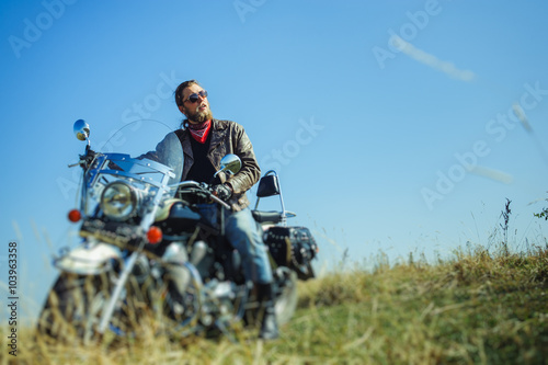 Portrait of a young man with beard sitting on his cruiser motorcycle. Biker is wearing leather jacket and blue jeans. Low point of view. Tilt shift lens blur effect